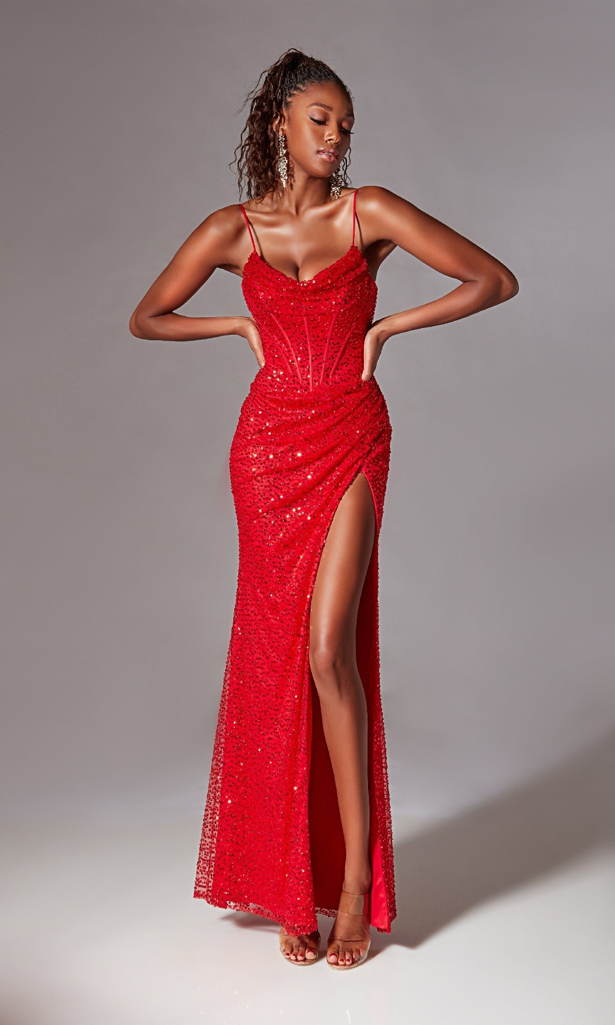 A woman wears the Corset Long Sequin Prom Dress: Aerie Couture 221—a fitted red gown with sequins, spaghetti straps, a corset-style bodice, and a high thigh slit—standing against a plain background.